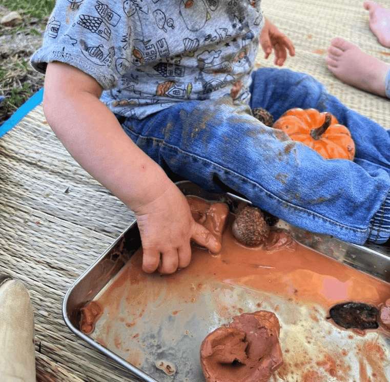 A child sits next to a tray with water and clay pieces